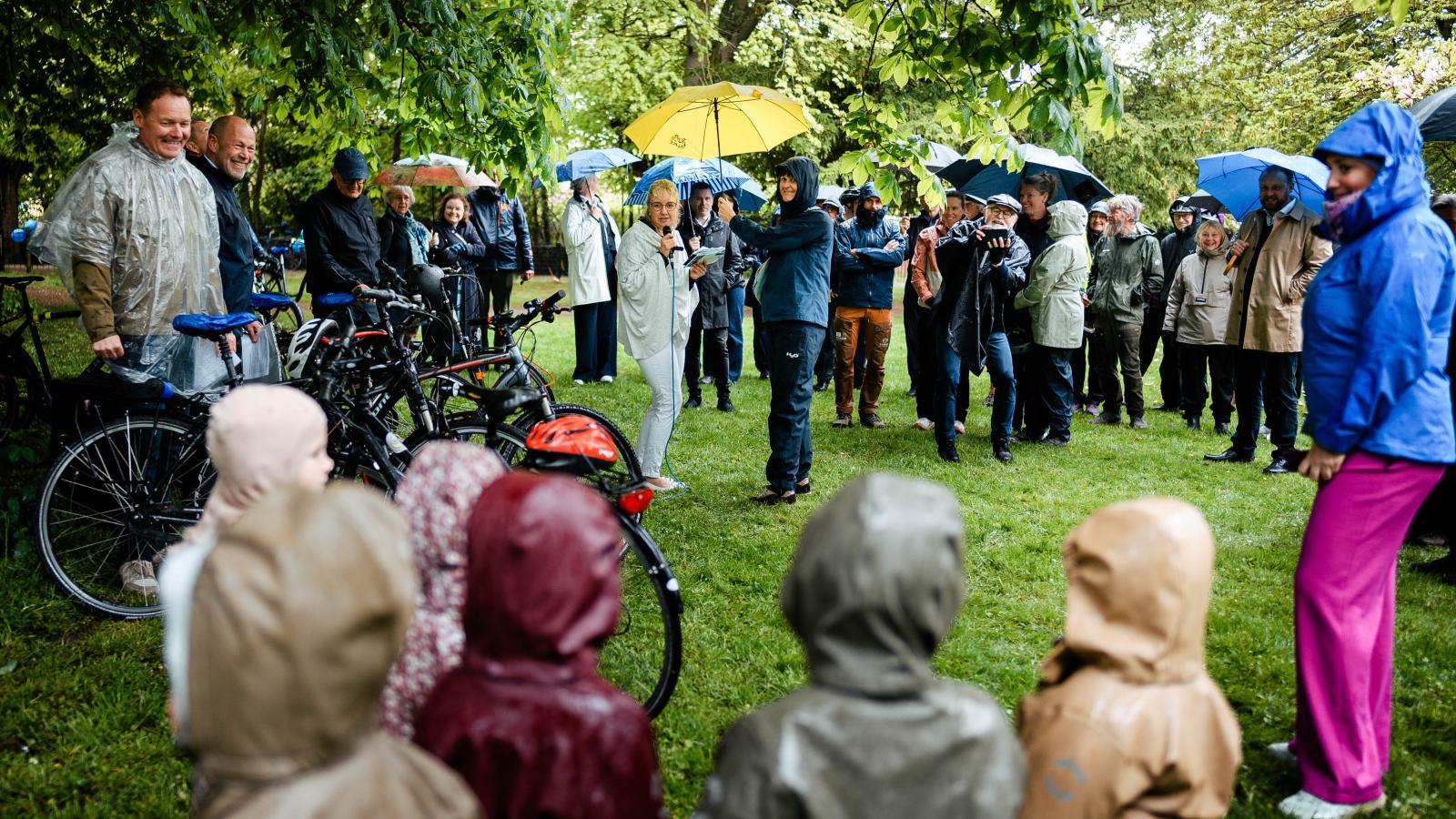 Forsamling i regntøj, regnslag og med paraplyer lytter til taler til lanceringen af Danmarks Rekreative Cykelnet