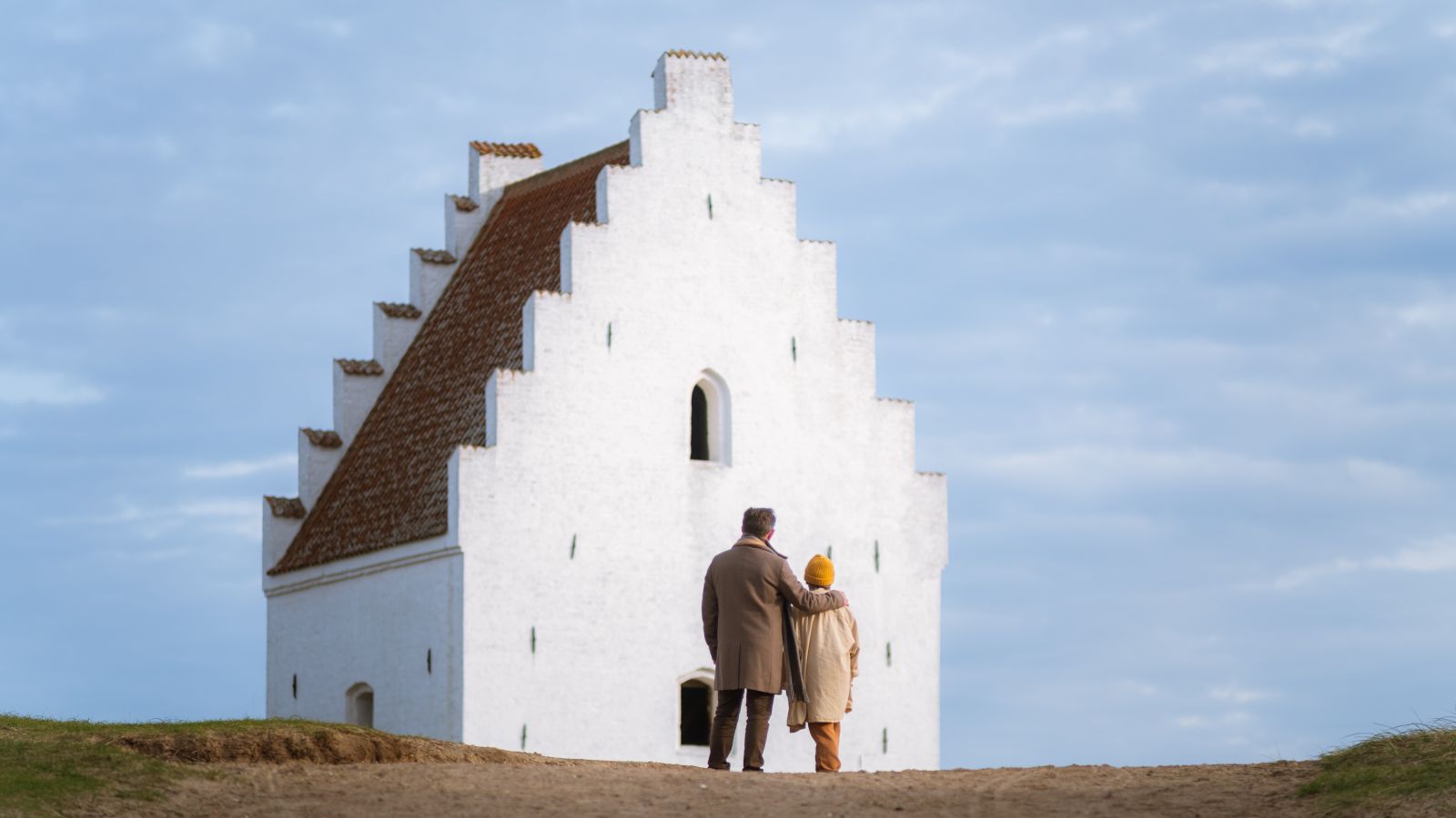 To personer står med ryggen til kameraet og kigger på den tilsandede kirke i Skagen