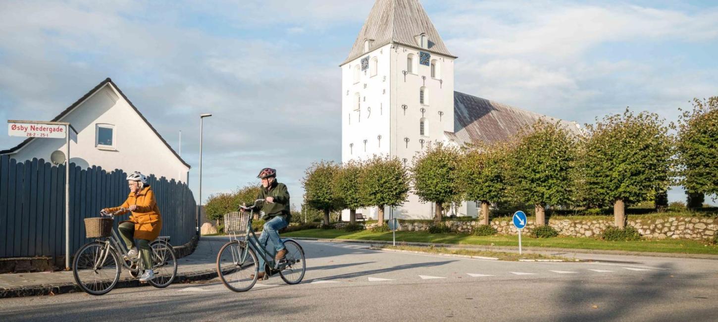 Et ældre par på cykeltur. I baggrunden kan man se en hvid kirke.