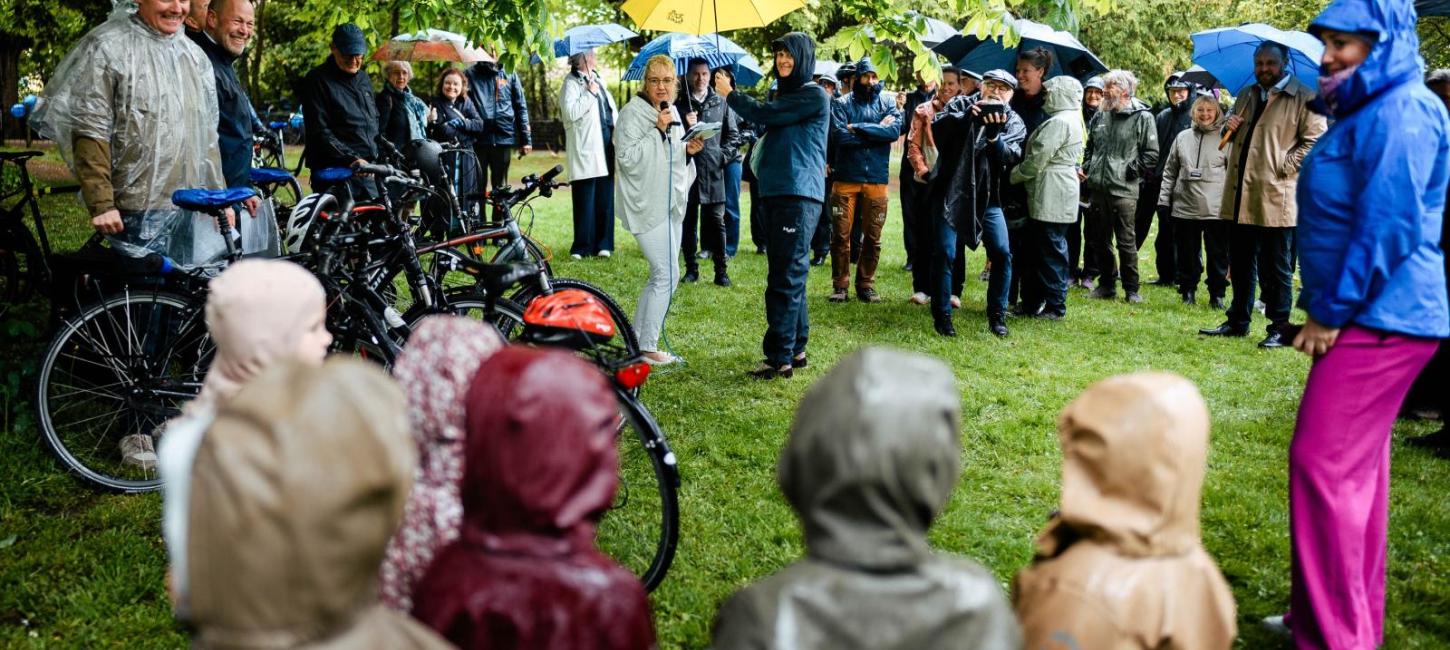 Forsamling i regntøj, regnslag og med paraplyer lytter til taler til lanceringen af Danmarks Rekreative Cykelnet